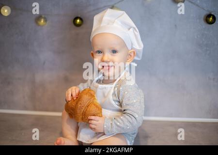 une jolie petite fille dans un chapeau de chef et un tablier est assise à la maison sur la table dans la cuisine avec un croissant Banque D'Images