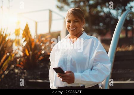 Un portrait en plein air d'une belle femme hispanique de taille plus mûre et gaie dans un t-shirt blanc et avec un smartphone dans la main, debout sur la str Banque D'Images