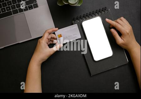 Une main féminine tenant une carte de crédit sur un bureau noir moderne avec une maquette d'écran blanc de smartphone pour afficher vos publicités graphiques, ordinateur portable et Banque D'Images