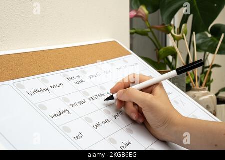 Femmes écrivant à la main avec un marqueur sur LE PLANIFICATEUR mensuel rempli de rendez-vous et de plans pour le mois prochain. Planning du mois chargé. Carte magnétique avec les jours du mois. Lieu pour entrer le calendrier des questions importantes. Concept de planification d'entreprise. Modèle mensuel magnétique du planificateur de tableau blanc. Intérieur du lieu de travail indépendant Banque D'Images