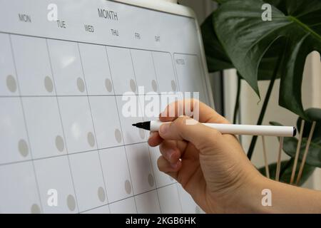 Femme écrivant à la main avec marqueur sur LE PLANIFICATEUR mensuel. Carte magnétique avec les jours du mois. Lieu pour entrer le calendrier des questions importantes. Concept de planification d'entreprise. Modèle mensuel magnétique du planificateur de tableau blanc. Intérieur du lieu de travail indépendant Banque D'Images