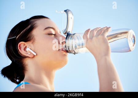 Eau potable, fitness et musique avec une femme de sport hydratant pendant une course sur fond de ciel bleu en plein air. Bien-être, santé et exercice avec un Banque D'Images