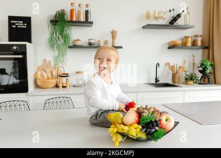 Un enfant heureux assis dans la cuisine souriant à côté de fruits et légumes. Banque D'Images
