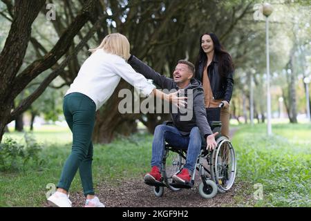 Les femmes amis marchant avec un homme handicapé en fauteuil roulant dans le parc, heureux de se voir Banque D'Images