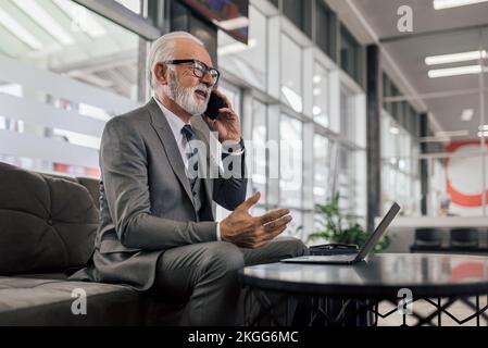 Un entrepreneur senior qui fait des gestes tout en parlant sur un téléphone intelligent. Un homme d'affaires âgé travaille sur un ordinateur portable au bureau. Banque D'Images