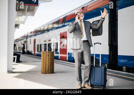 Souriant entrepreneur âgé qui fait des gestes tout en parlant sur un smartphone. Homme d'affaires est debout avec ses bagages à la plate-forme de la gare ou du métro. Il Banque D'Images