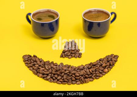 Émoticone souriant, lèvres en grains de café et yeux en tasses de café, composition sur fond jaune. Banque D'Images