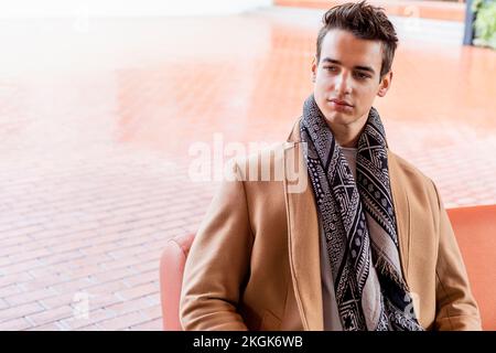 Jeune homme portant des vêtements d'hiver dans la rue. Jeune gars avec style moderne avec manteau, écharpe, jeans bleu et sweat blanc. Banque D'Images