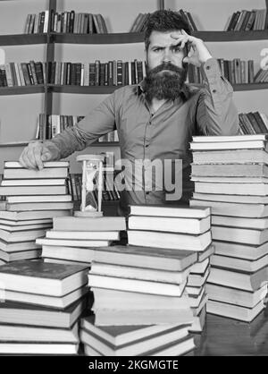 Concept de bibliothécaire. L'homme sur le visage réfléchi se tient entre des piles de livres, tout en étudiant dans la bibliothèque, les étagères sur le fond. Enseignant ou étudiant avec Banque D'Images