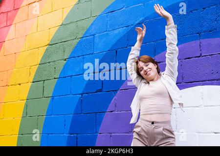 Femme heureuse avec les bras levés devant le drapeau arc-en-ciel peint sur le mur Banque D'Images