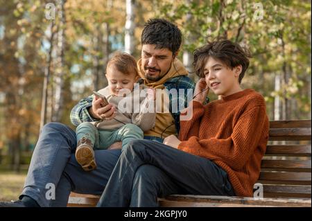 Homme avec son fils sur les genoux tenant un téléphone portable écoutant de la musique à travers des écouteurs intra-auriculaires assis par un garçon sur le banc Banque D'Images