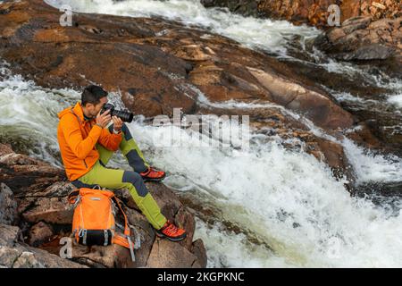 Homme mûr prenant des photos à travers l'appareil photo assis sur le rocher au bord de la rivière Banque D'Images