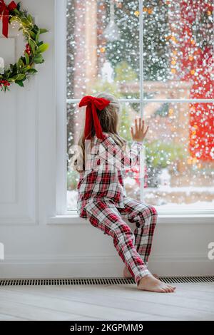 Fille en pyjama rouge regardant la neige à travers la fenêtre à la maison Banque D'Images