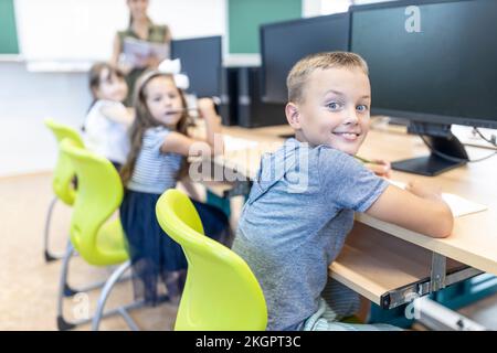 Garçon souriant regardant par-dessus l'épaule assis au bureau dans la salle de classe Banque D'Images