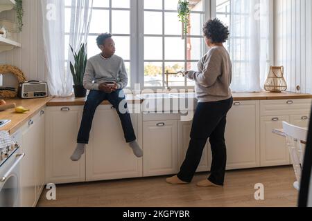 La mère lave les ustensiles avec son fils assis sur le comptoir de cuisine près de l'évier à la maison Banque D'Images