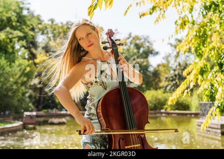 Femme blonde jouant du violoncelle devant le lac au parc Banque D'Images