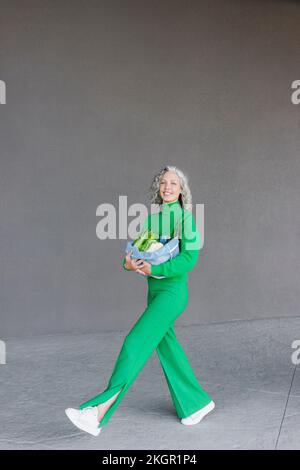 Femme souriante avec sac de légumes en filet marchant devant le mur Banque D'Images