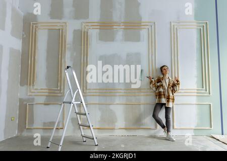 Femme souriante avec rouleau de peinture jour rêvant devant le mur à la maison Banque D'Images