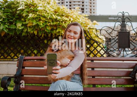 Bonne femme tenant un chien en train de prendre le selfie à travers un smartphone sur un banc dans le parc Banque D'Images