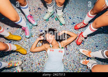Bonne jeune femme couché sur des confettis au milieu des jambes d'amis portant des patins à roulettes Banque D'Images