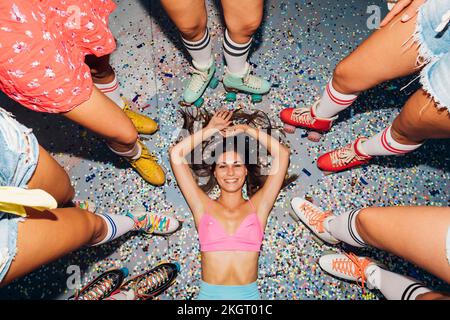 Femme couchée sur des confettis au milieu des jambes d'amis portant des patins à roulettes Banque D'Images