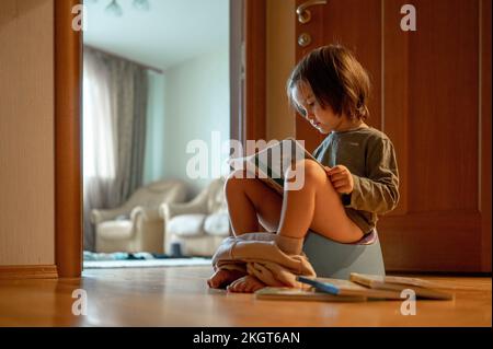 Mignon garçon assis sur pot et livre de lecture à la maison Banque D'Images