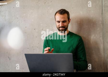 Homme d'affaires souriant effectuant des appels vidéo sur ordinateur portable, devant le mur au bureau Banque D'Images
