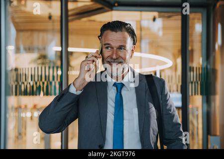 Homme d'affaires souriant et mûr parlant sur un smartphone devant une porte en verre Banque D'Images