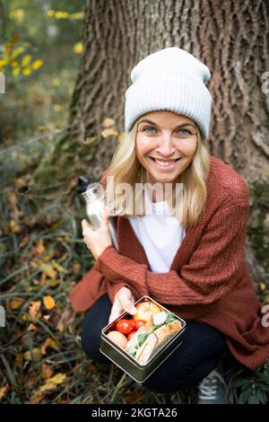 Femme souriante avec boîte à lunch qui s'accroupite dans la forêt le week-end Banque D'Images