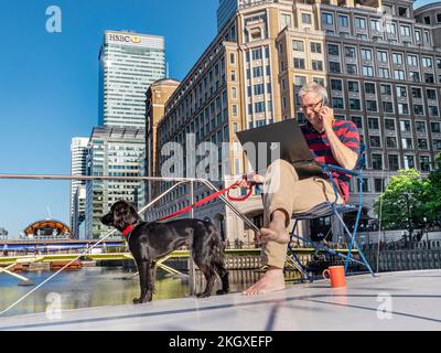 Big / Small Business décontracté homme adulte et chien d'animal assis à l'extérieur sur son bureau de péniche houseboat travaillant à son ordinateur portable Canary Wharf Londres E14 Banque D'Images