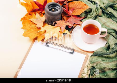 Bureau avec presse-papiers vierge, une tasse de thé, feuilles d'automne et bougie sur fond beige clair. Vue de dessus, maquette. Banque D'Images