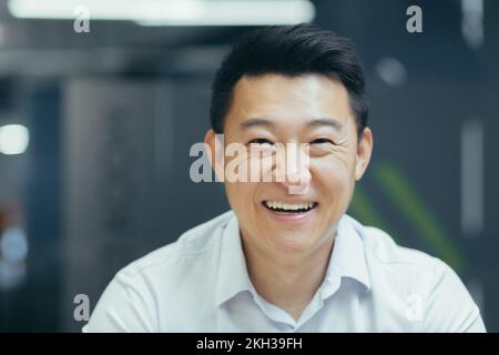 Gros plan. Portrait d'un beau jeune homme d'affaires asiatique. Il est assis au bureau dans une chemise blanche, regardant la caméra, souriant. Banque D'Images