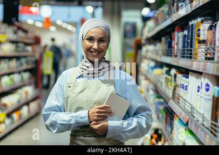 Portrait d'une femme dans un hijab, un ouvrier de magasin dans un supermarché avec une tablette, regarde la caméra et sourit, une femme vendeur consultant en lunettes parmi les rangées d'étagères avec des marchandises. Banque D'Images