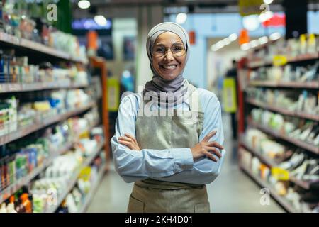 Portrait d'une vendeuse féminine dans un magasin de marchandises et d'étagères, une femme musulmane dans un tablier et un hijab est souriant et regarde la caméra, une vendeuse réussie. Banque D'Images