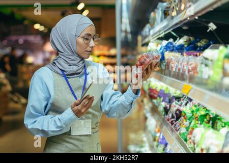 Une femme musulmane dans un hijab, un ouvrier de supermarché vérifie les produits sur les étagères et dans les réfrigérateurs, une femme avec une tablette et un tablier utilise une tablette pour examiner les produits. Banque D'Images