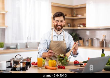 Un adulte souriant, un chef de cuisine européen barbu en tablier, prépare une salade dans un intérieur moderne Banque D'Images