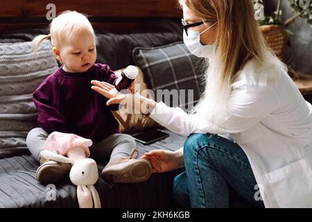 Petite fille blonde Cherubic assise sur un canapé, donnant une bouteille brune au médecin des enfants qui examine le patient à la maison. Banque D'Images