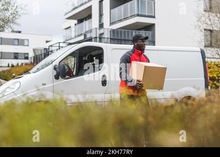 Ouvrier de col bleu noir - personne de livraison d'emballage - devant un camion blanc avec emballage en carton. Vêtements - veste rouge avec manches grises et chapeau noir. Herbe haute floue au premier plan. Photo de haute qualité Banque D'Images