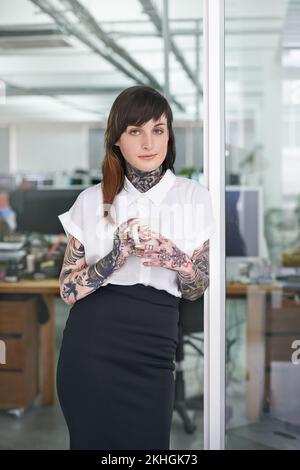 Stéréotypes - qui a besoin de em. Une photo courte d'une femme d'affaires tatouée debout dans son bureau avec une tasse de café. Banque D'Images