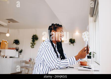 Femme d'affaires noire gaie ayant un appel vidéo sur un smartphone tout en travaillant dans un café. Bonne jeune femme d'affaires souriant lors d'un chat vidéo avec Banque D'Images