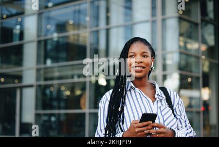 Femme d'affaires pensive tenant un smartphone et portant des écouteurs pendant ses déplacements professionnels en ville. Jeune femme d'affaires noire qui écoute de la musique Banque D'Images