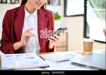 Image rognée, magnifique jeune femme d'affaires asiatique en costume d'affaires élégant en utilisant son smartphone à son bureau. chat, messagerie, internet. Banque D'Images