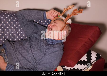 Homme de 50-60 ans dormant sur un canapé portant une décoration sous forme de cornes de cerf sur la tête. Homme reposant après Noël et fête du nouvel an. Banque D'Images