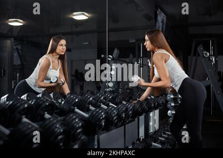 Mince, bodybuilder fille, soulève lourd haltère debout devant le miroir pendant l'entraînement dans la salle de gym. Concept sportif, combustion de graisse et une vie saine Banque D'Images