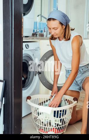 Une jeune femme de ménage est assise devant un lave-linge. Elle charge la machine à laver avec du linge sale. Banque D'Images