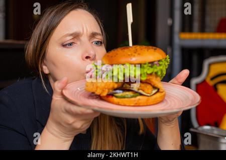 Drôle de tir femme soufflant et regardant un grand hamburger avec brochette sur l'assiette dans le restaurant. Bœuf, hamburger de porc avec petits pains Banque D'Images