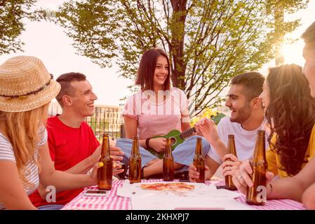 les jeunes du parc sont assis sur une table avec une nappe, une pizza et des bouteilles de bière, un groupe d'étudiants universitaires chantant, jouant et célébrant Banque D'Images