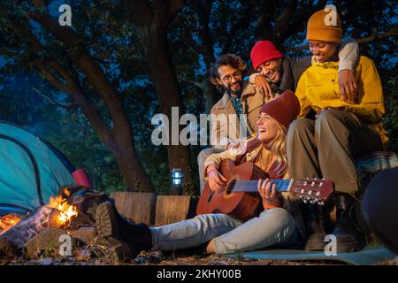 Des amis multiraciaux heureux au camping rient sur le feu tandis que la fille souriante joue de la guitare. Banque D'Images
