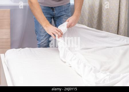 Un homme fait son lit confortable avec des draps blancs frais et lumineux. Faire le lit avec du linge de lit frais par un homme blanc. Le jour du changement de linge de lit. Le jour du lavage des linge de lit dans la buanderie. Banque D'Images