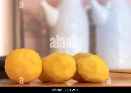 Trois pommes de terre, pelées et prêtes à cuire. Sur la planche à découper en bois. Outils de cuisine en arrière-plan. mise au point sélective. Banque D'Images